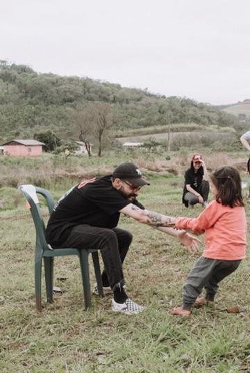 Matheus sentado numa cadeira no campo, brincando com uma menina
