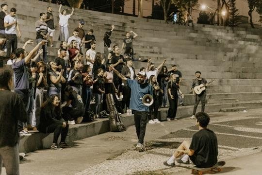 Culto na praça à noite, com Matheus ao megafone e jovens na escadaria